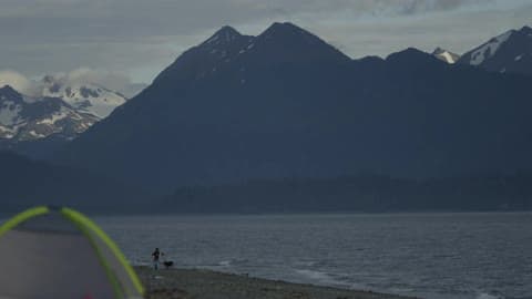 Family And Dog Stand Along Waters Edge On Beach With Snowy Mountains In Front Of Them