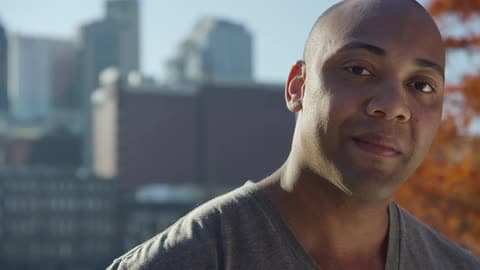 Man Smiles On A Sunny Day With A City Behind Him