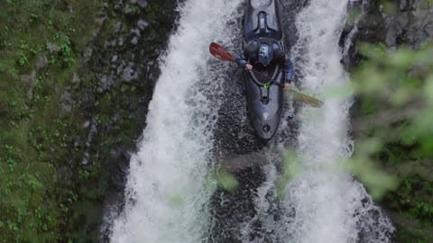 Tight Slow Motion Shot Of A Man In A Black Kayak Dropping A Huge Waterfall Deep In A Lush Green Canyon
